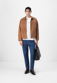 Brun mockajacka över vit t-shirt, blå jeans, svarta läderstövlar och bärande en svart väska. Enkelt mönster, slät textur.