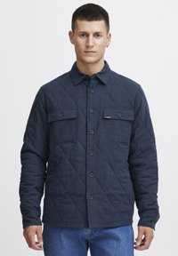 Dark blue quilted jacket with a button-up front, two chest pockets, and a collar. Made of soft fabric with a textured finish.