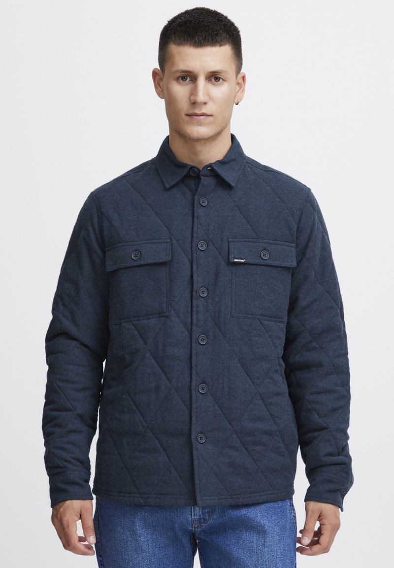 Dark blue quilted jacket with a button-up front, two chest pockets, and a collar. Made of soft fabric with a textured finish.