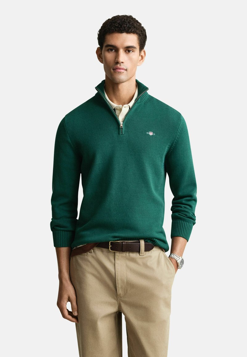 Grüner Pullover mit Reißverschlusskragen, gerippte Bündchen und kleinem Logo auf der Brust, getragen über einem hellen Hemd und beigen Hosen.