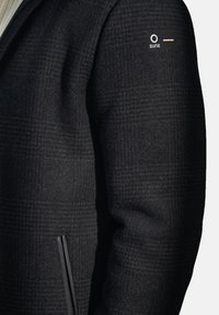 Manica di giacca a scacchi scuri con una tasca verticale nera con zip e un piccolo patch logo rettangolare sul braccio superiore.