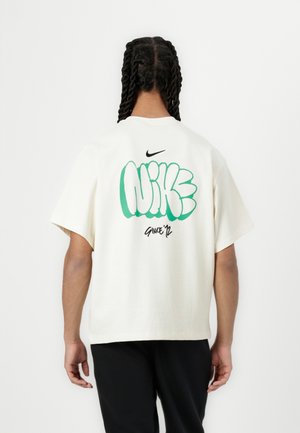 Osoba z plecionymi włosami, ubrana w białą koszulkę Nike z zielonym logo w stylu graffiti i czarne spodnie, stoi tyłem do widza.