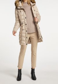 Beige donsjack met een bontvoering in de capuchon, rits- en knoopsluitingen, zijzakken, gedragen over een lichte trui en beige broek, gecombineerd met zwarte enkellaarsjes.