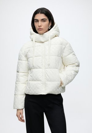 Frau trägt eine weiße gesteppte Kapuzenjacke mit dezentem Muster und schwarze Hose, steht mit einer Hand in der Tasche vor weißem Hintergrund.