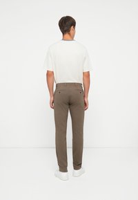 Brown cotton pants with a slim fit, featuring two back pockets and a smooth texture. The model wears a white t-shirt and white sneakers.