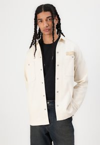 Vaaleanbeige napillinen takki, joka on tehty denimmateriilista ja jossa on kaksi etutaskua, hopeiset nepparit sekä teksturoitu merkki rinnassa. Takki on puettu mustan paidan päälle.