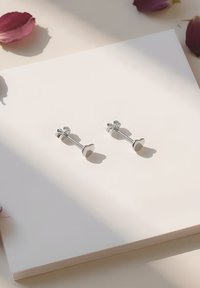 Silver-hjärtformade stiftörhängen som visas mot en ljus bakgrund med rosblad. Varje örhänge har en slät yta och en enkel design.