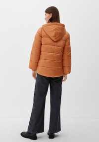 Kobieta widoczna z tyłu, ubrana w pomarańczową kurtkę puchową z kapturem, ciemne spodnie typu wide-leg i czarne buty, stojąca na jednolitym tle.