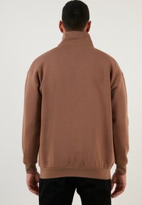 Su�éter marrón con cuello alto, fabricado en tejido suave. Presenta puños y bajo acanalados. Diseño sencillo con un corte relajado. Se muestra la vista trasera.