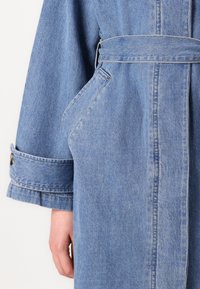 Giacca in denim azzurro chiaro con cintura in vita annodata, maniche lunghe e colletto a revers. Include tasche laterali e dettagli sui polsini.