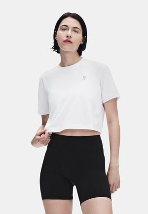 Kobieta o krótkich ciemnych włosach, ubrana w biały krótki t-shirt i czarne dopasowane krótkie spodenki z wysokim stanem na jednolitym tle.