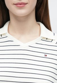 Krém színű, V-nyakú galléros póló tengerészkék vízszintes csíkokkal. Klasszikus gallérral és egy kis márkajelzéssel a bal mellrészen.