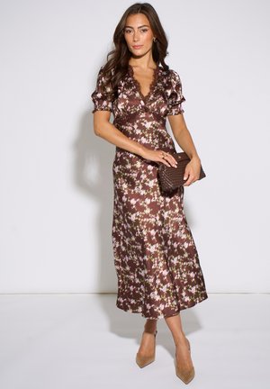 Woman wearing a brown floral midi dress with puff sleeves and holding a brown woven clutch, standing against a plain white background.
