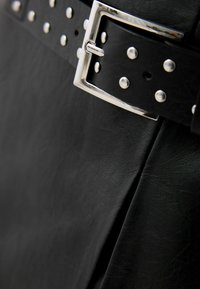 Cinturón de imitación de cuero negro con una hebilla de metal plateado y remaches. La textura es suave con un ligero brillo, destacando el diseño detallado.