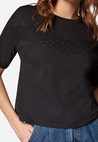 Černý bavlněný top s krátkým rukávem, který má texturovaný tkaný vzor přes hruď a malé výřezy, s mírně uvolněným střihem.