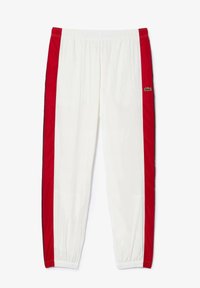Balti sportiniai kelnių su raudonomis šoninėmis juostelėmis. Elastinga juosmens juosta ir tankus raštas ties kojų galu. Žalias Lacoste logotipas ant raudonos šoninės juostos. Smoöka tekstūra.
