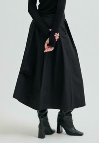 Osoba nosząca długą czarną spódnicę, czarną bluzkę z długim rękawem oraz czarne botki na obcasie, stojąca z rękami splecionymi za plecami.