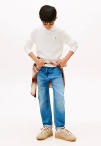 Chłopiec w białym swetrze, niebieskich dżinsach i beżowych trampkach, zawiązujący na talii koszulę w kratę, patrzący w dół.