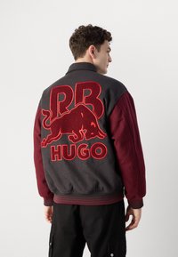 Varsity jacket in gray and burgundy with ribbed cuffs. Features large red embroidered "RB" and bull logo, and "HUGO" text on the back.