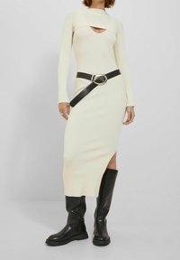 Kvinna klädd i en krämfärgad stickad midiklänning med ett nyckelhålshalssmycke, svart oversized bälte och svarta knähöga l äderstövlar.