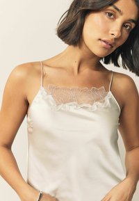 Silk camisole in off-white with thin straps and a floral lace trim at the neckline, creating a delicate texture and detail.