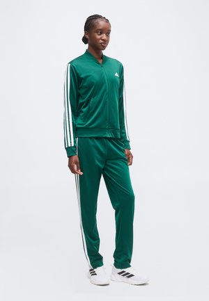 Młoda kobieta ubrana w zielony dres Adidas z białymi paskami i białe buty sportowe Adidas, stojąca na jednolitym tle.