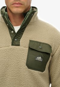 Beige fleece sweatshirt med en grøn krave med trykknapper og en brystlomme. Har et broderet logo og en struktureret overflade.