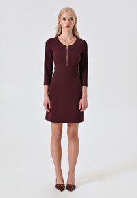 Abito aderente color malva con maniche a tre quarti, scollo rotondo e zip frontale. Accentuato con un tira zip dorato e una texture liscia.