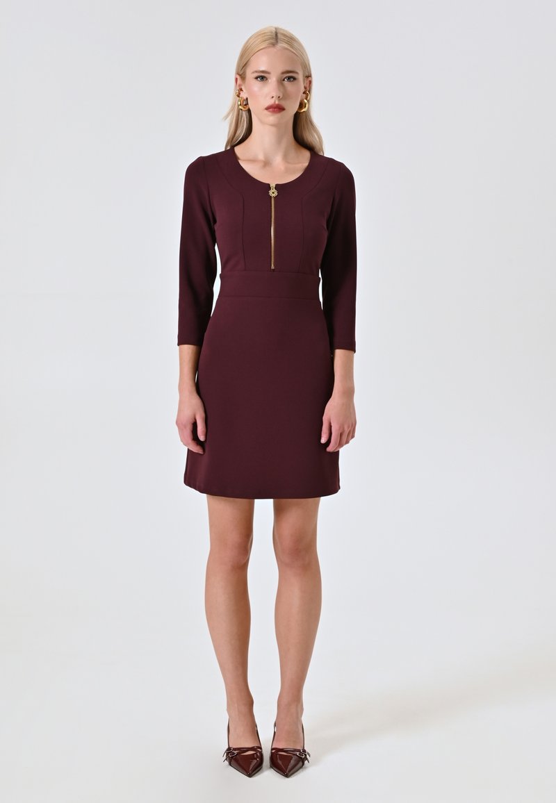 Abito aderente color malva con maniche a tre quarti, scollo rotondo e zip frontale. Accentuato con un tira zip dorato e una texture liscia.