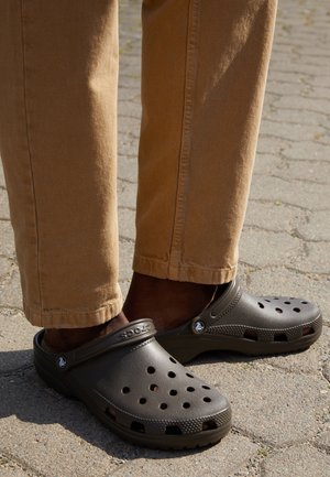 Dark brown Crocs clogs with ventilation holes, textured sole, and a back strap, paired with light brown, slightly tapered pants.