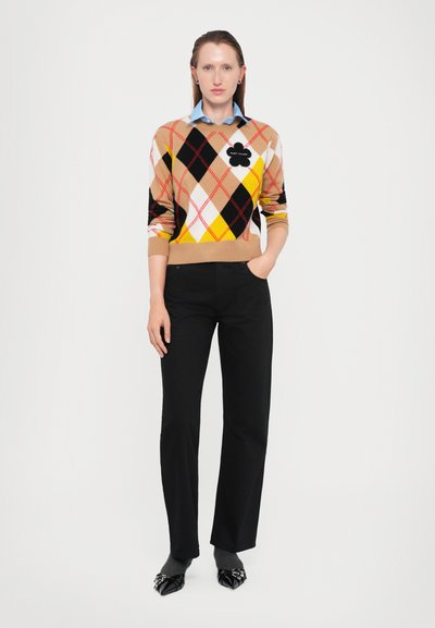 Sweater with argyle pattern in tan, black, yellow, and white; features a black floral logo. Paired with black straight-leg trousers and shoes.
