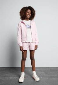 Felpa leggera rosa con zip e tasche, pantaloni corti rosa abbinati e una maglietta bianca con grafica. Sneakers alte bianche con accenti rossi.