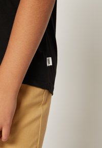 Camiseta de algodón negra de manga corta, con una etiqueta blanca de Vans en el dobladillo, combinada con pantalones de textura color marrón claro.