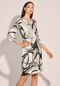 Schwarz-weiß bedrucktes Kleid mit dreiviertel Ärmeln, das wirbelnde Linien und eine Knotendetail an der Taille zeigt, aus strukturiertem Stoff gefertigt.