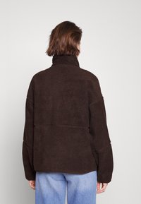 Brązowa polarowa kurtka z wysokim kołnierzem, w oversize'owym kroju, z opadającymi ramionami i teksturowaną powierzchnią. Połączona z jasnoniebieskimi dżinsami.