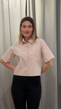 Γυναίκα με μαλλιά μέχρι τους ώμους φοράει ανοιχτό ροζ κοντομάνικο πουκάμισο με κουμπιά και μαύρο παντελόνι, στέκεται με τα χέρια πίσω από την πλάτη απέναντι σε ανοιχτή κουρτίνα.