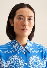 Camicia blu con colletto, caratterizzata da motivi paisley bianchi e righe. Il tessuto sembra liscio e presenta una chiusura con bottoni.