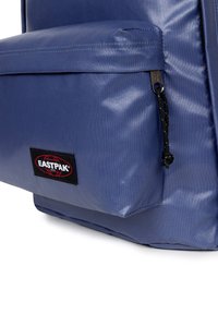 Niebieski plecak z tkaniny o gładkiej strukturze, z przednią kieszenią zamykaną na czarny zamek oraz czerwono-czarną naszywką z logo.