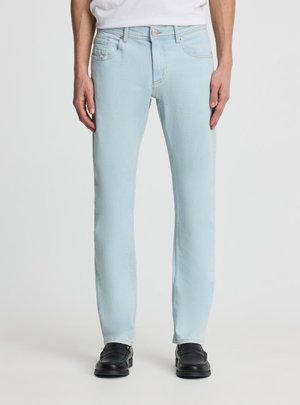 Jeans slim fit azzurri chiari indossati da una persona in piedi, abbinati a mocassini neri e a una camicia bianca infilata.