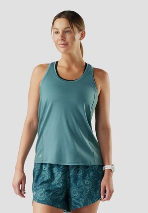 Donna con canotta sportiva turchese e pantaloncini con motivo floreale, in piedi contro uno sfondo semplice, che indossa un orologio da polso bianco.