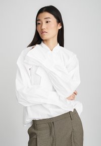 Camicia bianca a maniche lunghe con colletto alto e dettaglio a fiocco sulle maniche, abbinata a pantaloni marroni con texture. Design semplice e pulito.