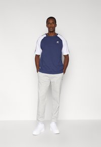 Κοντομάνικο μπλουζάκι σε ναυτικό μπλε με λευκές λεπτομέρειες και λογότυπο, σε συνδυασμό με ανοιχτογκρι φόρμα και λευκά αθλητικά παπούτσια. Το μοντέλο στέκεται με τα χέρια στις τσέπες.