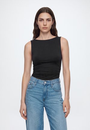 Mujer con pelo largo y castaño que lleva una camiseta negra sin mangas y vaqueros azules de talle alto, de pie frente a un fondo blanco liso.