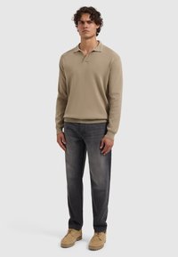 Mann steht mit einem beigen langärmeligen Hemd-Pullover, ausgewaschenen schwarzen Jeans und beigen Schnürschuhen vor einem schlichten hellen Hintergrund.