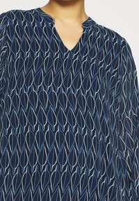 Marine-blaues Blusenshirt mit V-Ausschnitt, das ein sich wiederholendes Blattmuster mit hellblauen Konturen aufweist. Hergestellt aus glattem, leichtem Stoff.