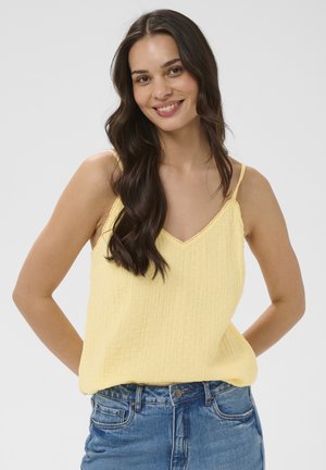 Femme souriante aux longs cheveux foncés, portant un haut jaune sans manches et un jean bleu, debout, les mains dans le dos.