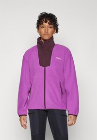 Purple fleece jacket with a brown high collar, featuring two zippered side pockets and contrasting fabric panels. Logo on the chest.