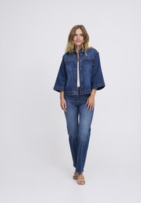 Denimjack met een cropped ontwerp, twee voorkwesten en knoopsluiting. Draag het over een witte top met blauwe rechte jeans.