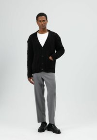 Zwarte kabelgebreide cardigan met een diepe V-hals, knopen aan de voorkant en zakken, gecombineerd met lichtgrijze broek en zwarte loafers.