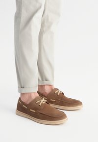 Hnědé semišové boty s béžovými šňůrkami a švy, spárované se světle béžovými nohavicemi s ohrnutím, které ukazují volný design.